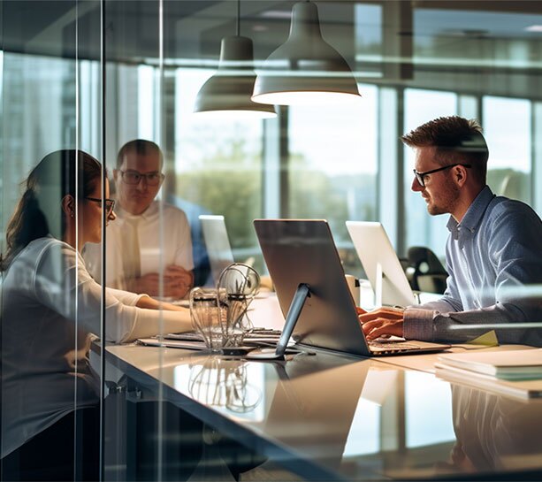 Business professionals working on laptops in a glass-walled office, collaborating in a modern, open workspace with natural light.