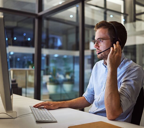Customer support agent wearing a headset and glasses, speaking while working at a computer in a modern office environment.